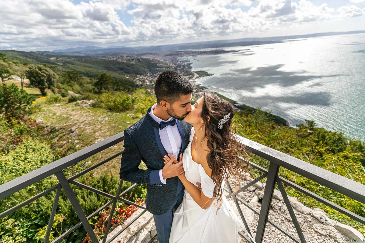 Coppia di sposi si bacia su una terrazza con vista panoramica sul mare e il paesaggio circostante.